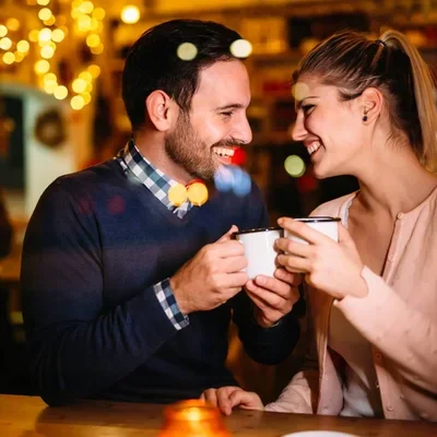 a man and sitting on the left and a woman sitting on the right, smiling at each other with cups in their hands.